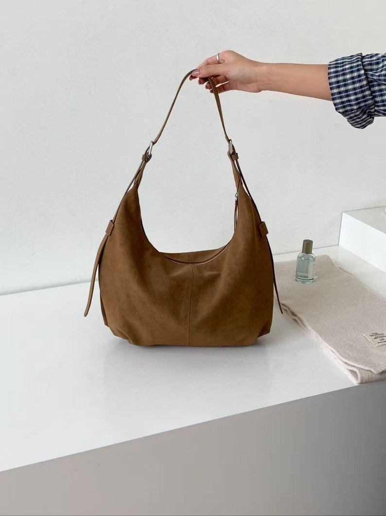 May include: A brown suede shoulder bag with a curved design. The bag has a long strap and silver-colored hardware. The bag is on a white surface. A hand is holding the strap. A small perfume bottle and a folded scarf are also on the surface.
