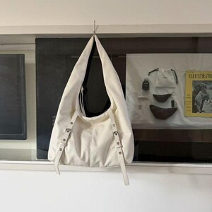 Puede incluir: Bolso de hombro color crema con un dise&ntilde;o holgado, colgado de un gancho de metal. El bolso presenta correas ajustables con herrajes plateados. El interior del bolso es negro. El bolso est&aacute; hecho de un material texturizado.