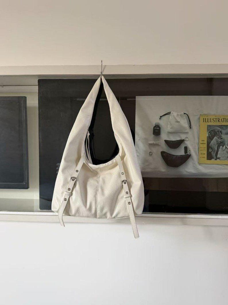 Puede incluir: Bolso de hombro color crema con un dise&ntilde;o holgado, colgado de un gancho de metal. El bolso presenta correas ajustables con herrajes plateados. El interior del bolso es negro. El bolso est&aacute; hecho de un material texturizado.