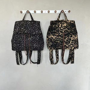 May include: Two leopard print backpacks hanging on a white wall. The backpacks have a black and tan spotted pattern, with adjustable straps and top handles. The bags are made of a soft fabric.