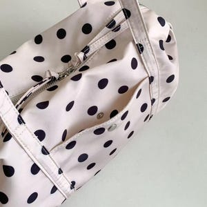 Puede incluir: Una bolsa de lona rosa claro con lunares negros. La bolsa tiene un compartimento principal con cremallera, un bolsillo frontal con cierre de bot&oacute;n a presi&oacute;n y dos asas. La bolsa es de material liso, posiblemente nailon.