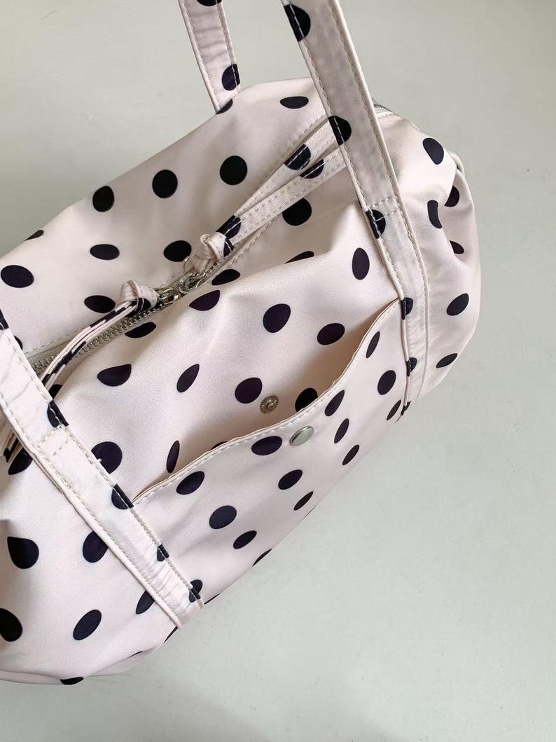 Puede incluir: Una bolsa de lona rosa claro con lunares negros. La bolsa tiene un compartimento principal con cremallera, un bolsillo frontal con cierre de bot&oacute;n a presi&oacute;n y dos asas. La bolsa es de material liso, posiblemente nailon.