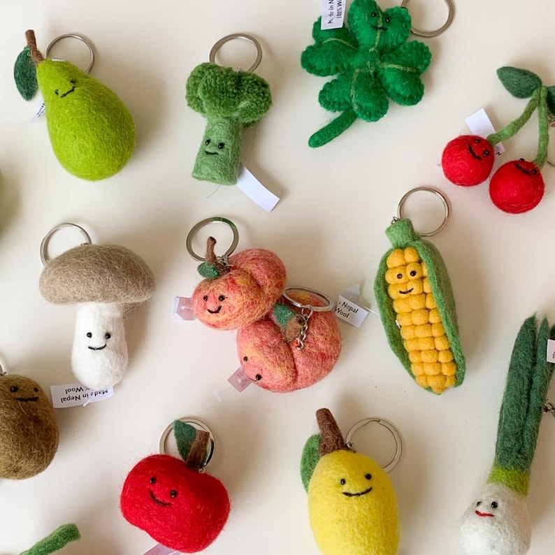 May include: A collection of felt food keychains, including a green broccoli, a yellow corn on the cob, a red apple, a green pear, a brown mushroom, a yellow lemon, a red cherry, a green clover, and a peach. The keychains are made in Nepal and are made of wool.