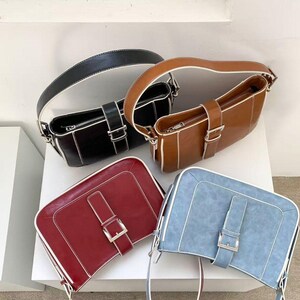 May include: Four rectangular handbags in black, brown, red, and blue. Each bag has a matching strap, a silver buckle, and white stitching along the edges. The bags are displayed on a white surface.