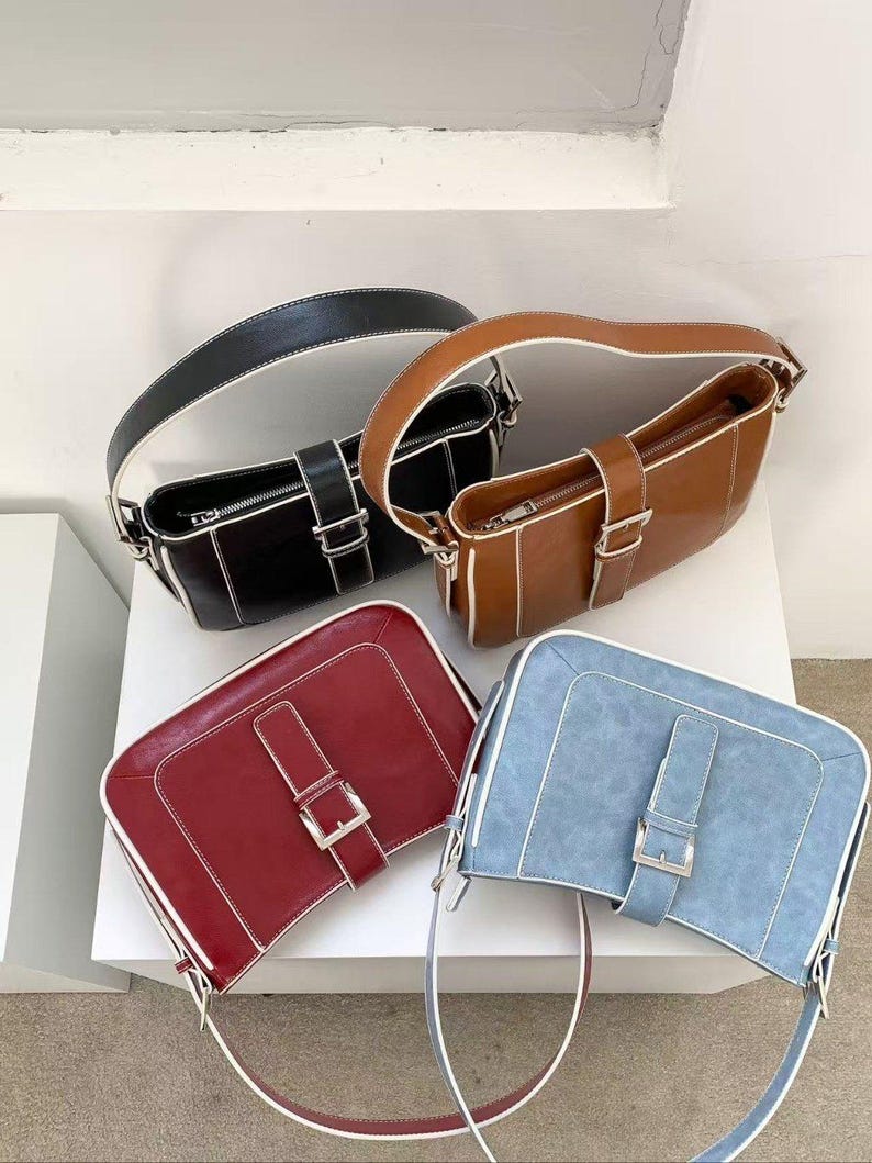 May include: Four rectangular handbags in black, brown, red, and blue. Each bag has a matching strap, a silver buckle, and white stitching along the edges. The bags are displayed on a white surface.