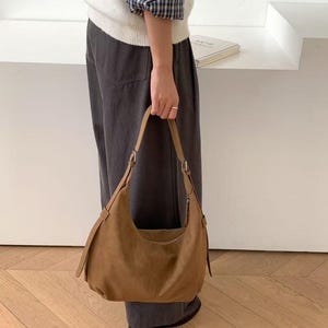 May include: A brown suede shoulder bag with a curved design. The bag has a long strap and a zipper closure. The bag is held by a person wearing a white sweater and dark gray pants.
