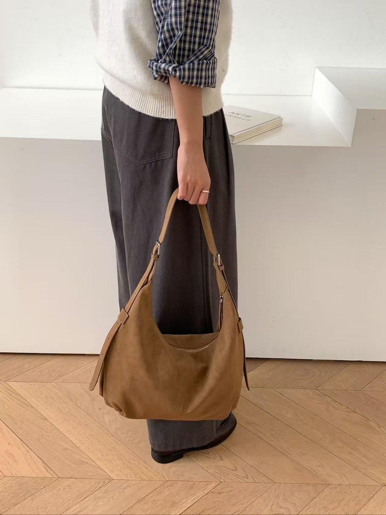 May include: A brown suede shoulder bag with a curved design. The bag has a long strap and a zipper closure. The bag is held by a person wearing a white sweater and dark gray pants.