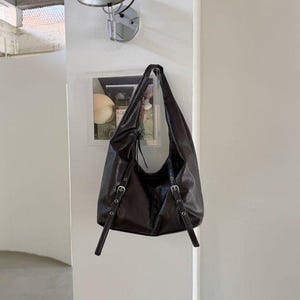 Puede incluir: Un bolso de hombro de cuero marr&oacute;n oscuro con correas ajustables y hebillas plateadas. El bolso est&aacute; colgado de una pared. El bolso tiene un dise&ntilde;o holgado y un solo compartimento.