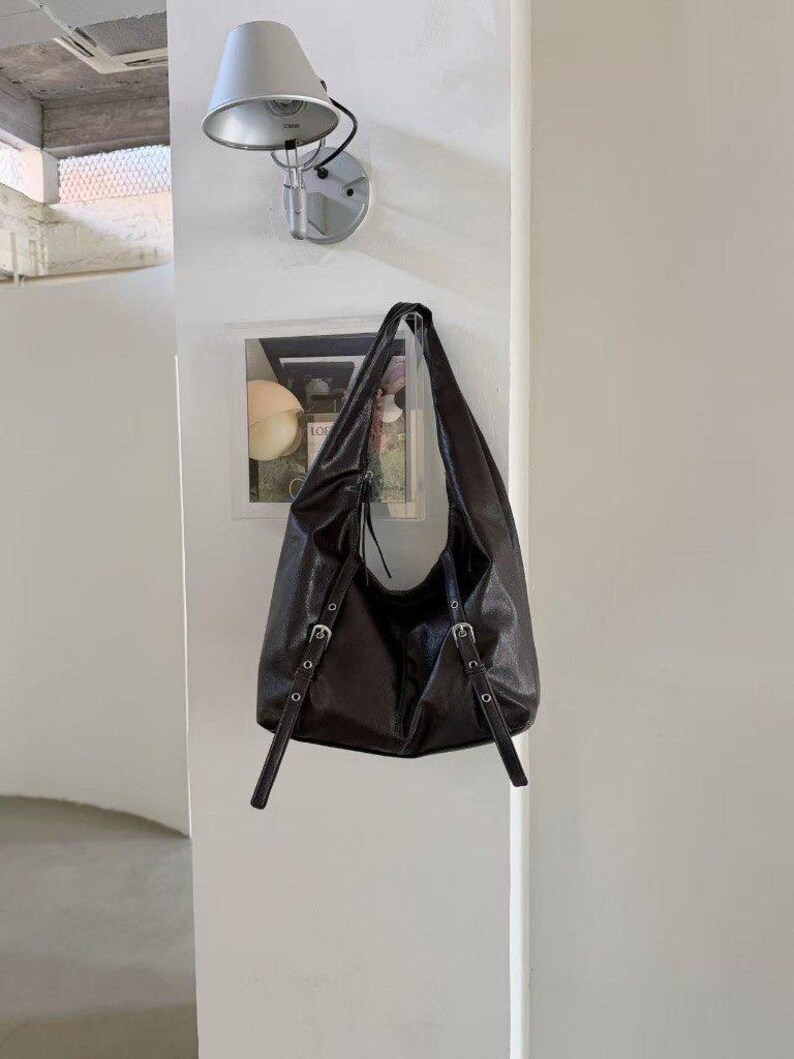 Puede incluir: Un bolso de hombro de cuero marr&oacute;n oscuro con correas ajustables y hebillas plateadas. El bolso est&aacute; colgado de una pared. El bolso tiene un dise&ntilde;o holgado y un solo compartimento.