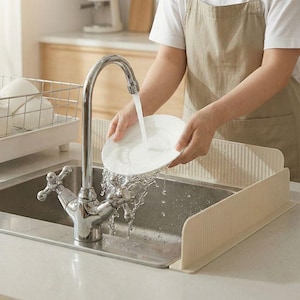 May include: A person washes a white plate under a chrome faucet in a stainless steel sink. Beige sink splash guards are installed on either side of the sink. A white dish rack is visible in the background.