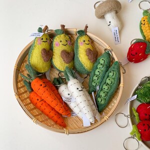 May include: A collection of felt food keychains in a bamboo basket. The keychains include avocados, carrots, radishes, peas, and strawberries. Each keychain has a smiling face and is made of 100% wool. The tag reads "Made in Nepal 100% Wool".