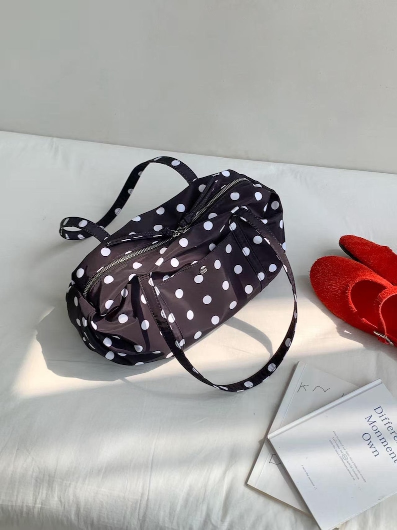 Puede incluir: Bolso de viaje negro con lunares blancos, con cierre de cremallera y dos asas. La bolsa est&aacute; hecha de un material ligero. Zapatos rojos y libros en el fondo.