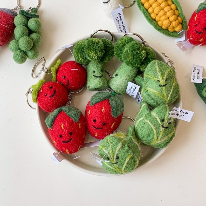 May include: A collection of felt food keychains, including strawberries, broccoli, and lettuce, all with happy faces. The keychains are made of 100% wool and are made in Nepal.