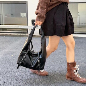 Puede incluir: Bolso de hombro de cuero negro con herrajes plateados. El bolso tiene forma curva y correas largas. La persona lleva un jersey marr&oacute;n, pantalones cortos negros y botas marrones.