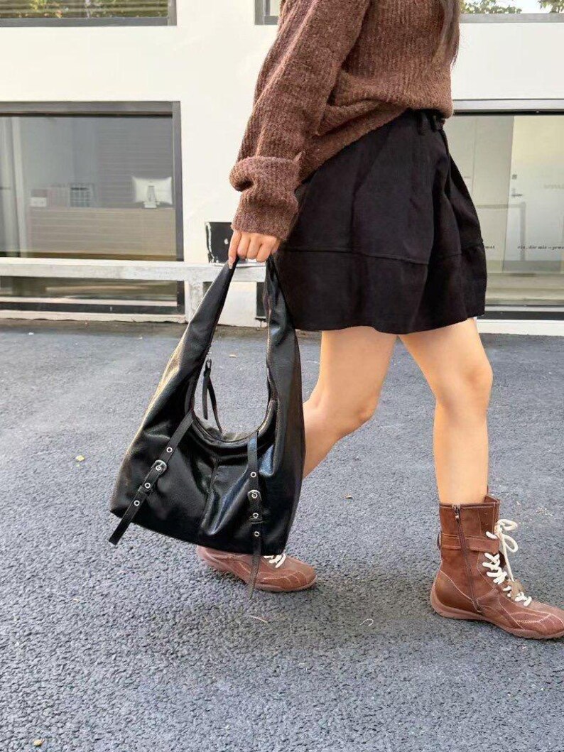 Puede incluir: Bolso de hombro de cuero negro con herrajes plateados. El bolso tiene forma curva y correas largas. La persona lleva un jersey marr&oacute;n, pantalones cortos negros y botas marrones.