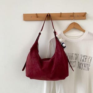 May include: A burgundy suede shoulder bag hangs from a wooden wall hook. The bag has a curved shape with a long strap. A white t-shirt with the text "EXCEL NOTHING" is visible on a hanger behind the bag.