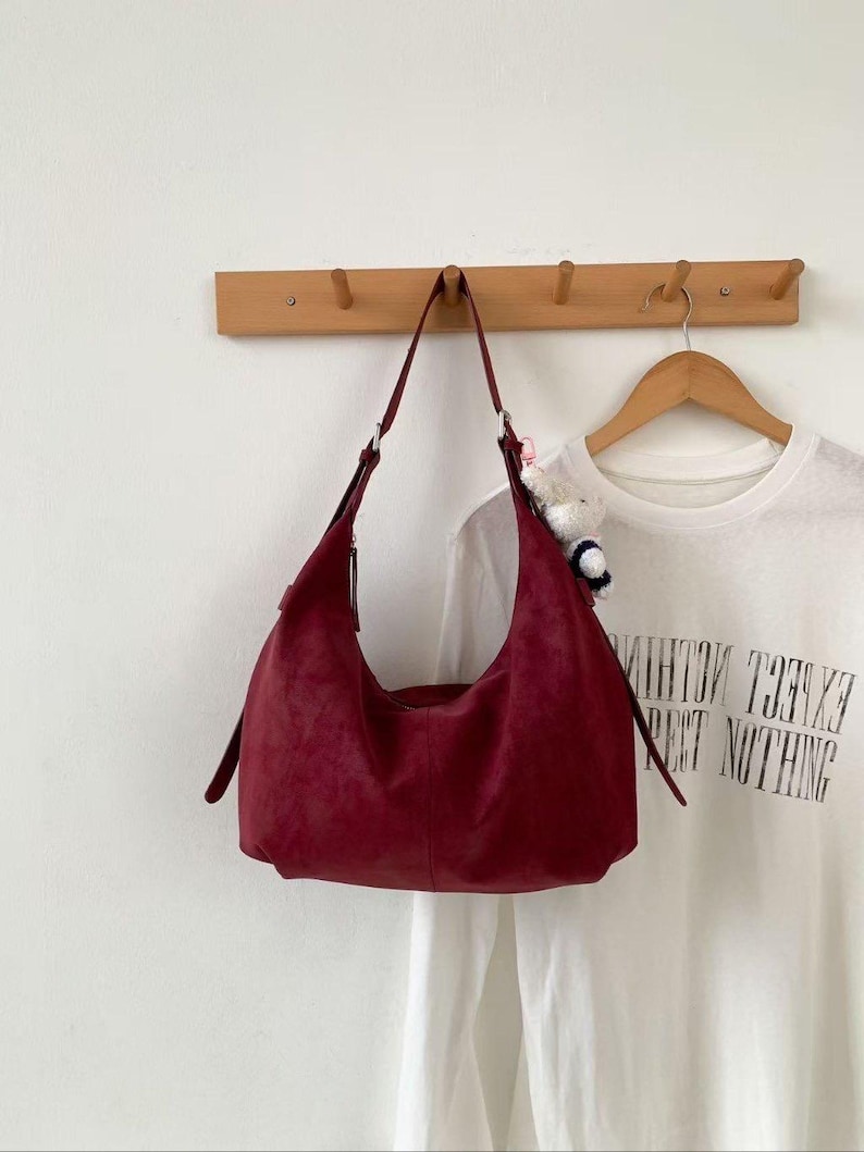 May include: A burgundy suede shoulder bag hangs from a wooden wall hook. The bag has a curved shape with a long strap. A white t-shirt with the text "EXCEL NOTHING" is visible on a hanger behind the bag.