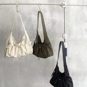 May include: Three shoulder bags in cream, olive green, and black hang from a metal bar. Each bag has a long strap and a gathered, ruched design at the bottom. The bags are made of a soft, possibly nylon, material.