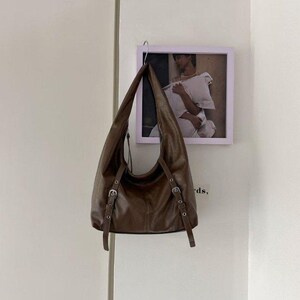 Puede incluir: Un bolso de hombro de cuero marr&oacute;n con correas ajustables y hebillas plateadas cuelga de un gancho. El bolso tiene una forma curva y un dise&ntilde;o suave y holgado. Una foto enmarcada cuelga de la pared detr&aacute;s del bolso.