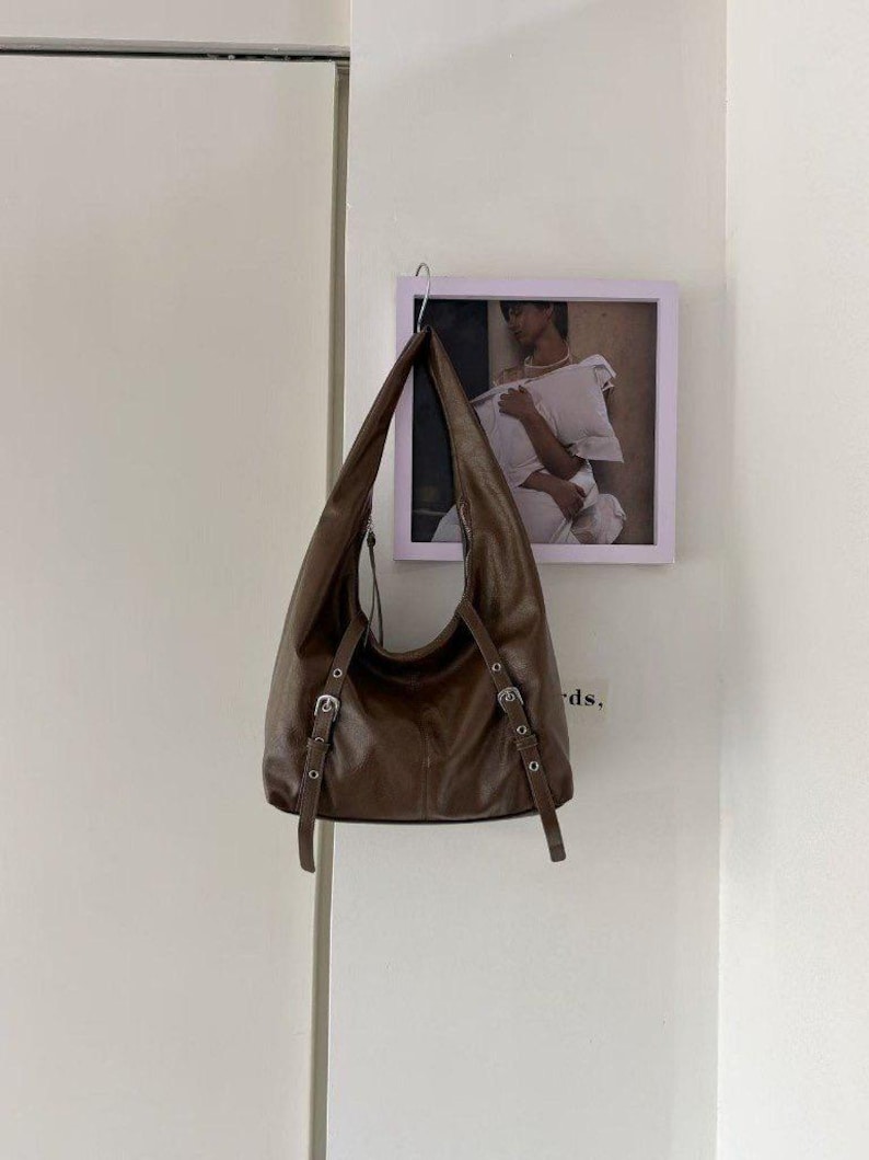 Puede incluir: Un bolso de hombro de cuero marr&oacute;n con correas ajustables y hebillas plateadas cuelga de un gancho. El bolso tiene una forma curva y un dise&ntilde;o suave y holgado. Una foto enmarcada cuelga de la pared detr&aacute;s del bolso.