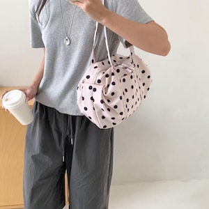 Puede incluir: Un bolso de mano rosa claro con un estampado de lunares negros. El bolso tiene forma redondeada, cierre de cremallera y correa para el hombro. Una persona lleva una camiseta gris y pantalones grises oscuros.