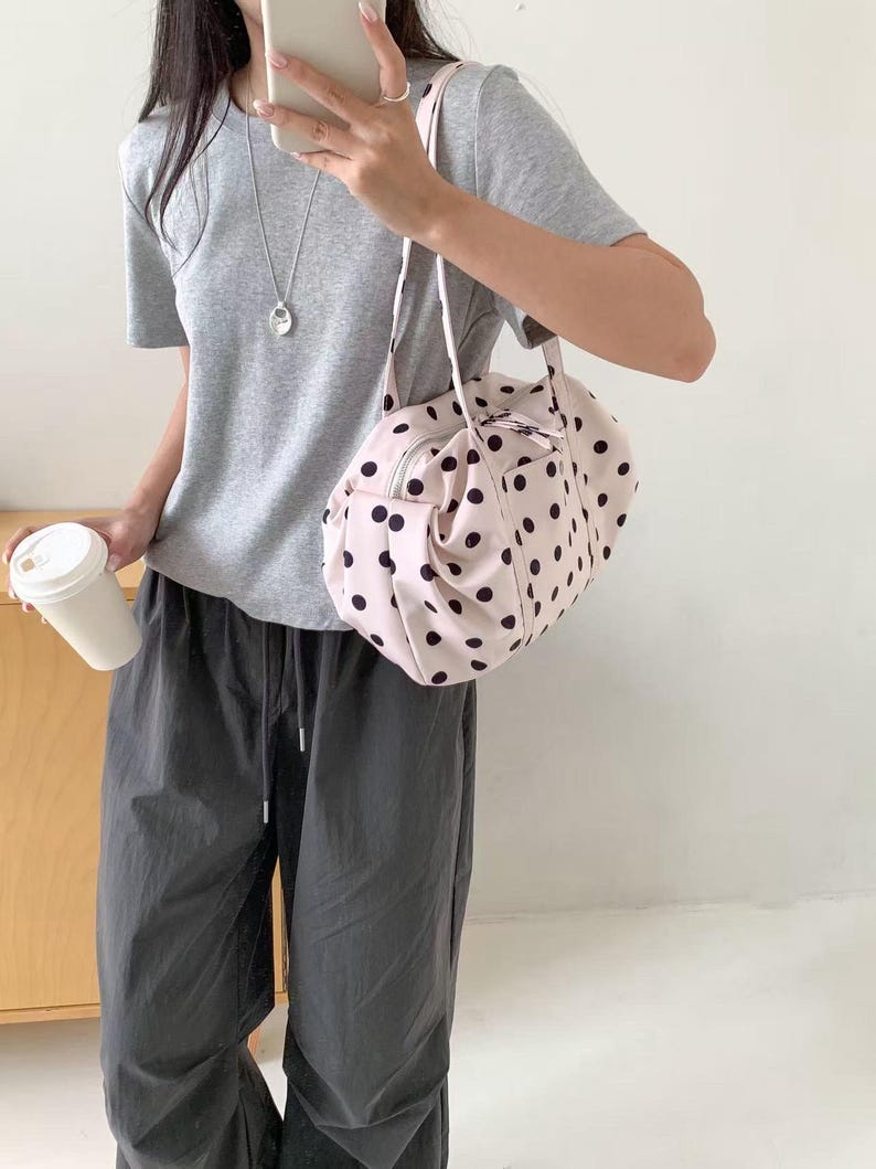 Puede incluir: Un bolso de mano rosa claro con un estampado de lunares negros. El bolso tiene forma redondeada, cierre de cremallera y correa para el hombro. Una persona lleva una camiseta gris y pantalones grises oscuros.