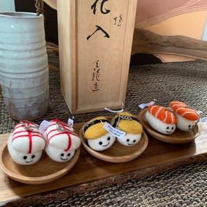 May include: A wooden tray displays felt sushi keyrings with smiling faces. The sushi pieces include red and white, yellow and black, and orange and white designs. A wooden box with Japanese characters and a ceramic vase are in the background.