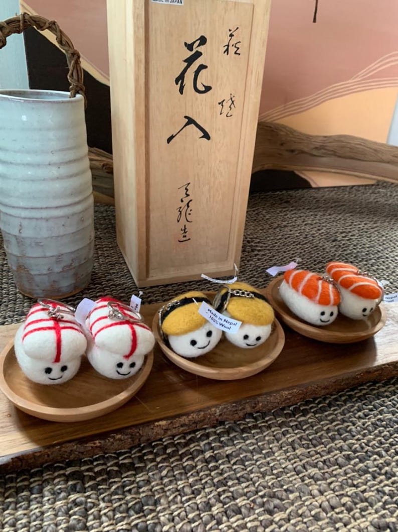 May include: A wooden tray displays felt sushi keyrings with smiling faces. The sushi pieces include red and white, yellow and black, and orange and white designs. A wooden box with Japanese characters and a ceramic vase are in the background.