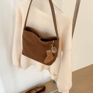 May include: A brown suede shoulder bag with a matching strap and a small boot-shaped charm. The bag is hanging on a clothing rack with a cream-colored sweater. A pair of brown flats are on a shelf below.