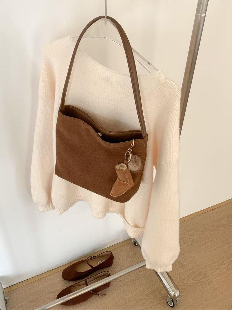 May include: A brown suede shoulder bag with a matching strap and a small boot-shaped charm. The bag is hanging on a clothing rack with a cream-colored sweater. A pair of brown flats are on a shelf below.