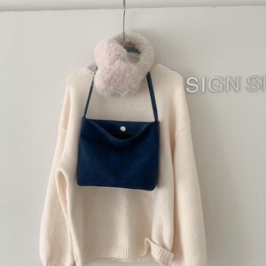 May include: A cream-colored sweater with a navy blue crossbody bag and a fluffy pink scarf hanging on a metal hook. The bag has a button closure. The sweater has a ribbed hem and cuffs.