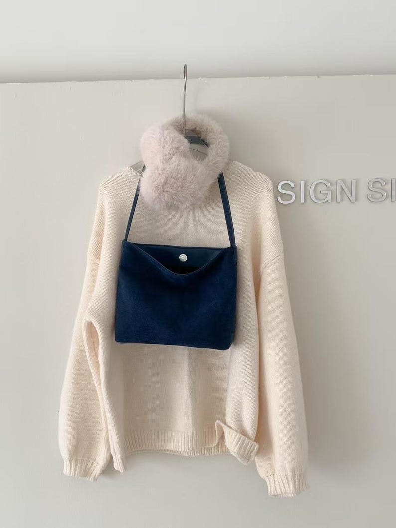 May include: A cream-colored sweater with a navy blue crossbody bag and a fluffy pink scarf hanging on a metal hook. The bag has a button closure. The sweater has a ribbed hem and cuffs.