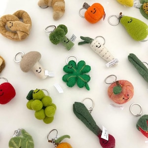 May include: A selection of felt keyrings in the shapes of food and plants, such as a pretzel, broccoli, mushroom, grapes, clover, pumpkin, pear, carrot, apple, and radish. Each keyring has a metal ring and a smiling face.