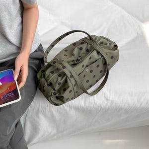 Puede incluir: Una bolsa de lona verde oliva con un patr&oacute;n repetido de lunares morados oscuros. La bolsa tiene dos asas y un bolsillo con cremallera en la parte delantera. La bolsa est&aacute; sobre una superficie blanca.