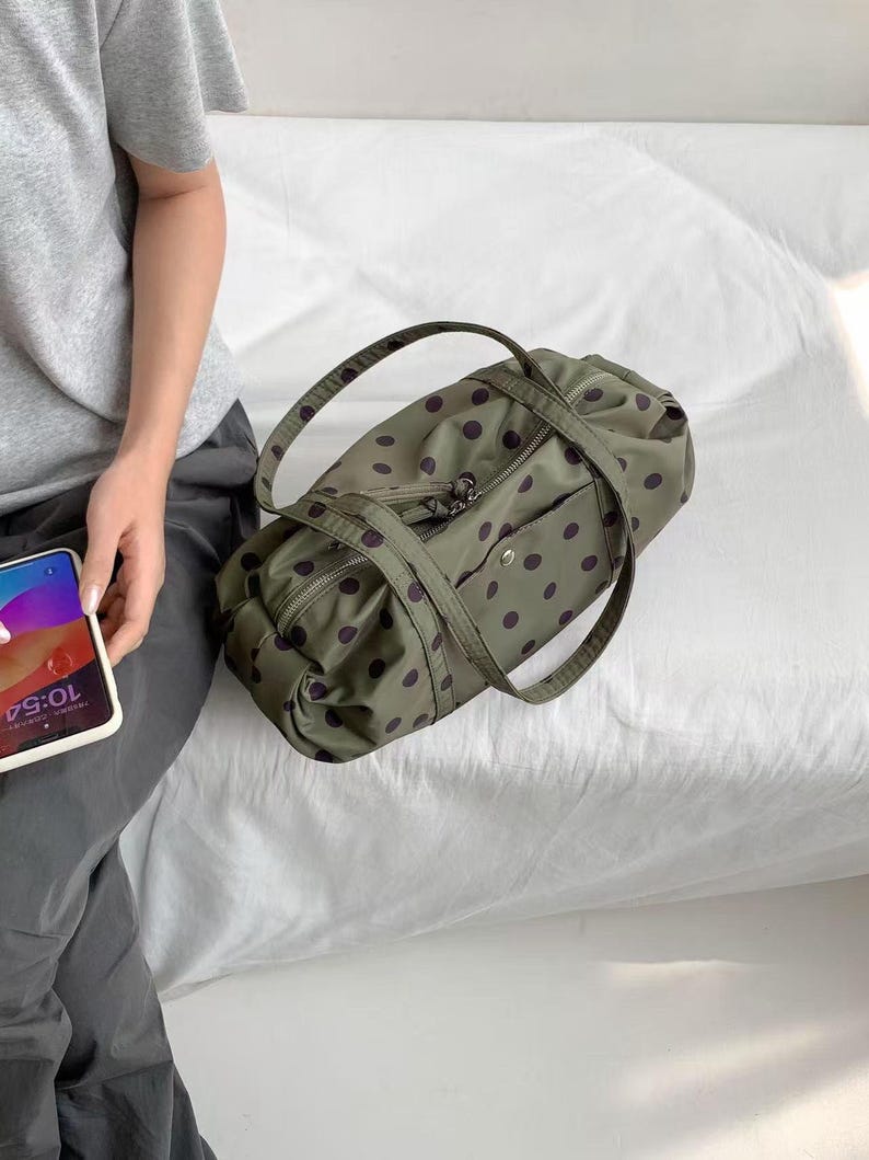 Puede incluir: Una bolsa de lona verde oliva con un patr&oacute;n repetido de lunares morados oscuros. La bolsa tiene dos asas y un bolsillo con cremallera en la parte delantera. La bolsa est&aacute; sobre una superficie blanca.