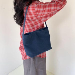 May include: A navy blue shoulder bag with a rectangular shape. The bag has a long strap and is worn over the shoulder. The person is wearing a red plaid shirt and gray pants.