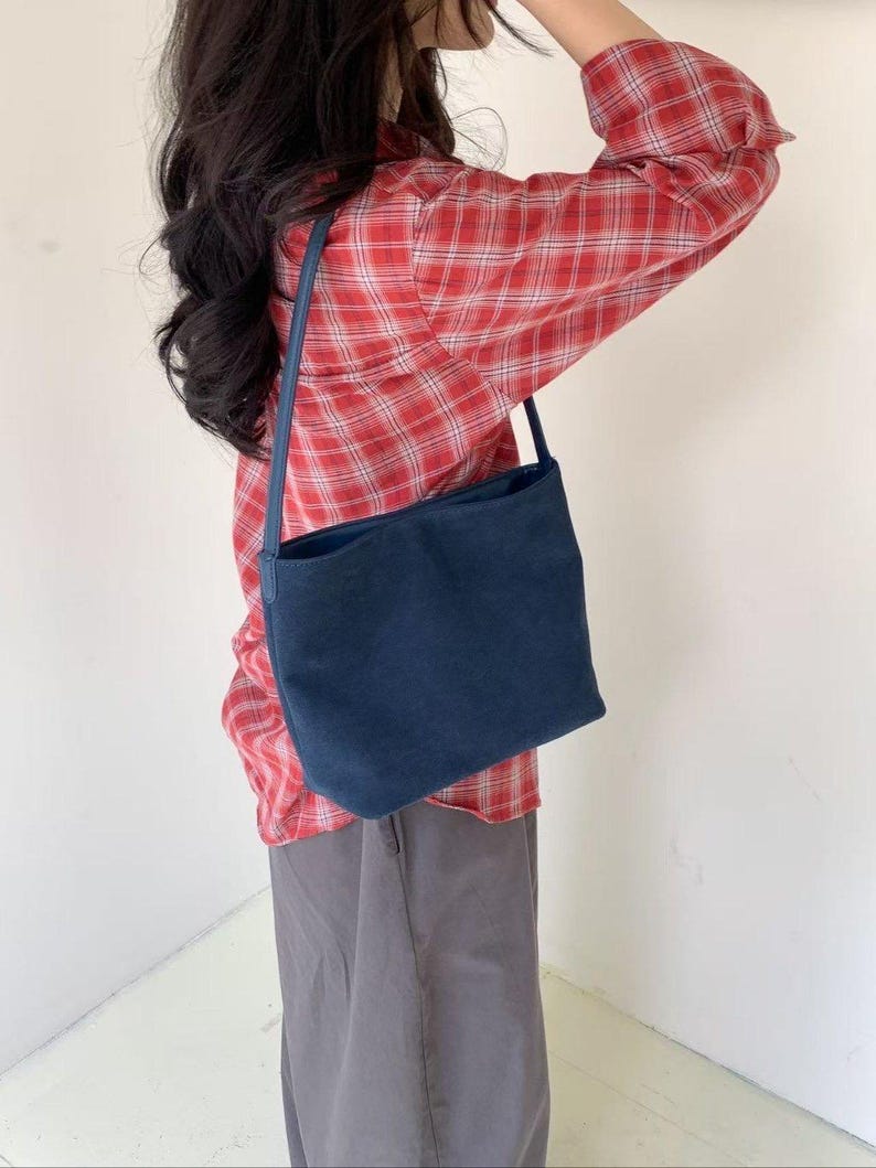 May include: A navy blue shoulder bag with a rectangular shape. The bag has a long strap and is worn over the shoulder. The person is wearing a red plaid shirt and gray pants.