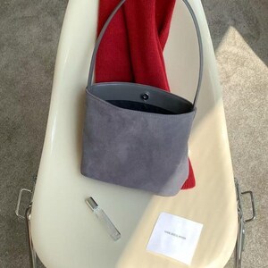 May include: A gray suede handbag with a single handle sits on a white chair. A red scarf is draped over the chair's back. A small perfume bottle and a white card with text are also on the chair.