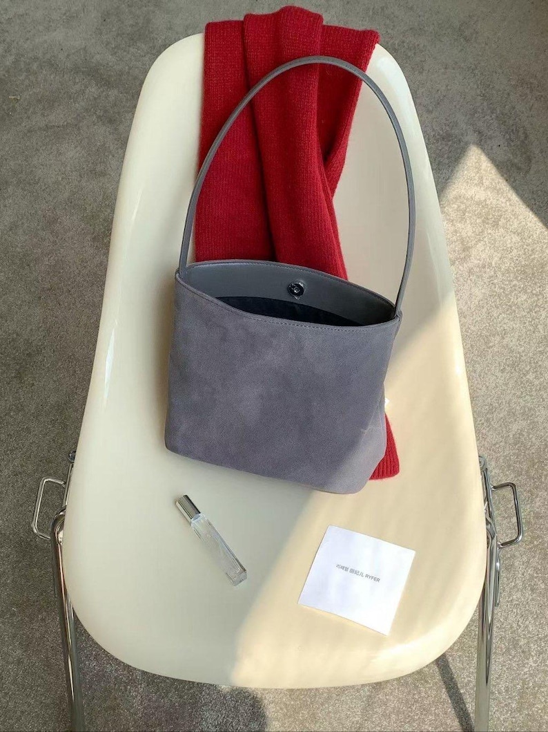 May include: A gray suede handbag with a single handle sits on a white chair. A red scarf is draped over the chair's back. A small perfume bottle and a white card with text are also on the chair.