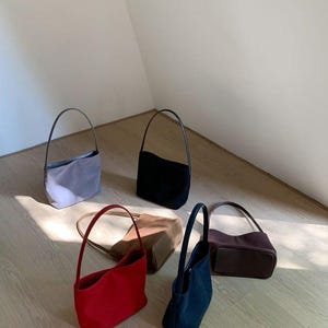 May include: An assortment of small, suede-like handbags in various colors, including red, black, navy, brown, and gray. Each bag has a slender, curved handle. The bags are arranged on a light-colored wooden floor.