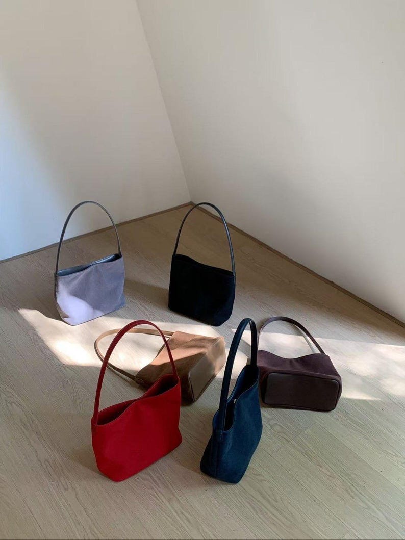 May include: An assortment of small, suede-like handbags in various colors, including red, black, navy, brown, and gray. Each bag has a slender, curved handle. The bags are arranged on a light-colored wooden floor.