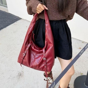 Puede incluir: Un bolso de hombro de cuero burdeos con correas ajustables y hebillas plateadas. El bolso tiene un dise&ntilde;o grande y abierto y un brillo sutil. El bolso lo sostiene una persona que lleva un su&eacute;ter marr&oacute;n y pantalones cortos negros.