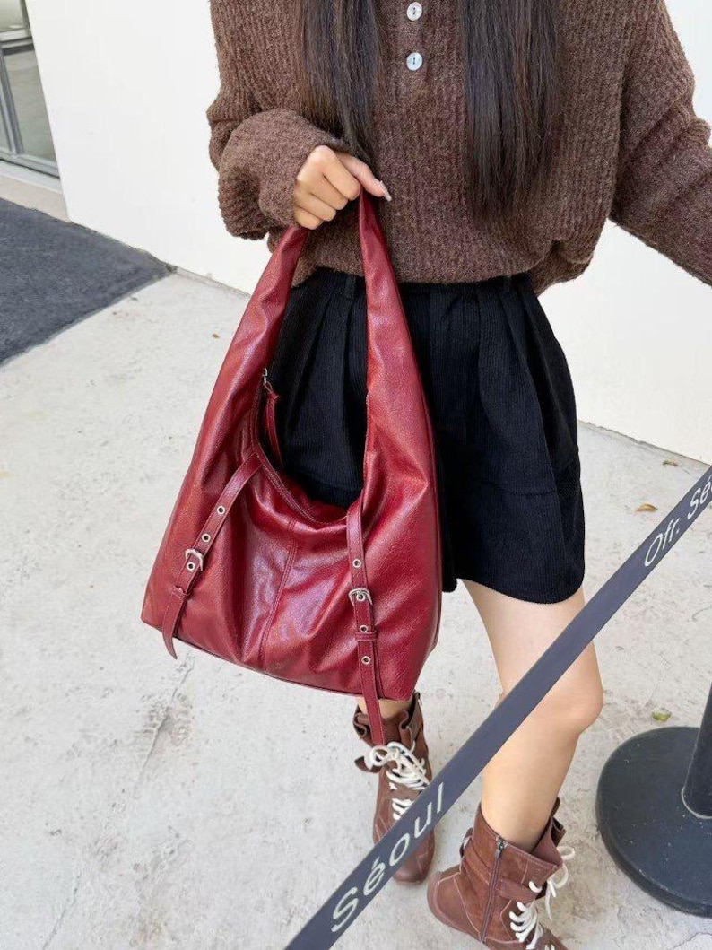 Puede incluir: Un bolso de hombro de cuero burdeos con correas ajustables y hebillas plateadas. El bolso tiene un dise&ntilde;o grande y abierto y un brillo sutil. El bolso lo sostiene una persona que lleva un su&eacute;ter marr&oacute;n y pantalones cortos negros.