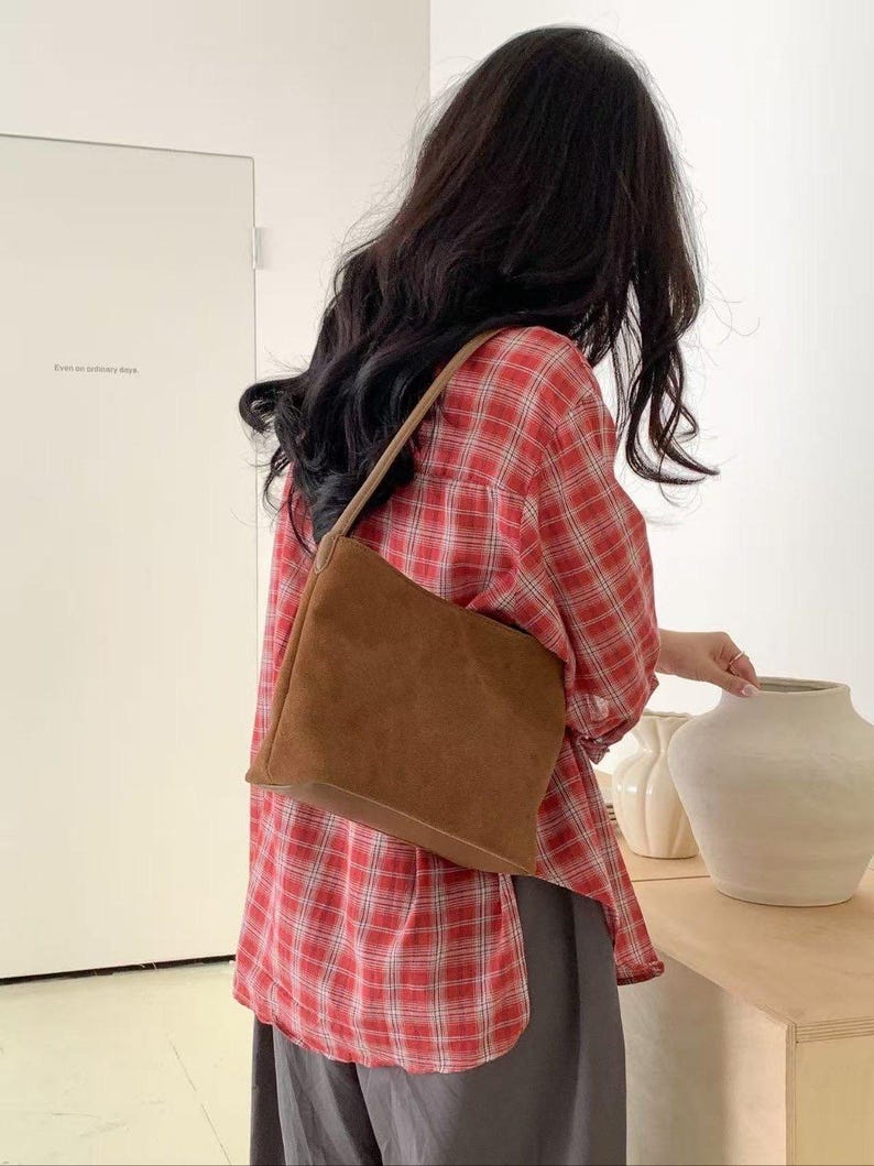 May include: A brown suede shoulder bag with a rectangular shape and a thin strap. The bag is worn over a red and white plaid shirt and gray pants. The background includes a white door and decorative vases.