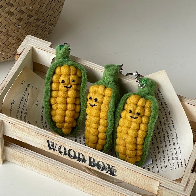 May include: Three felt corn on the cob keychains with happy faces. The corn is yellow and the husks are green. The keychains are in a wooden crate.