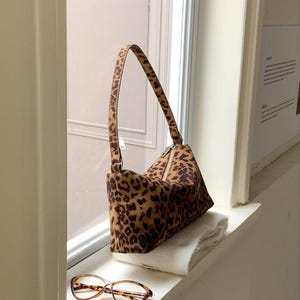 以下が含まれることがあります： 茶色とベージュのヒョウ柄ハンドバッグ。ジッパー開閉と、お揃いのストラップ付き。べっ甲柄の眼鏡が、バッグの下の白い面に置かれています。