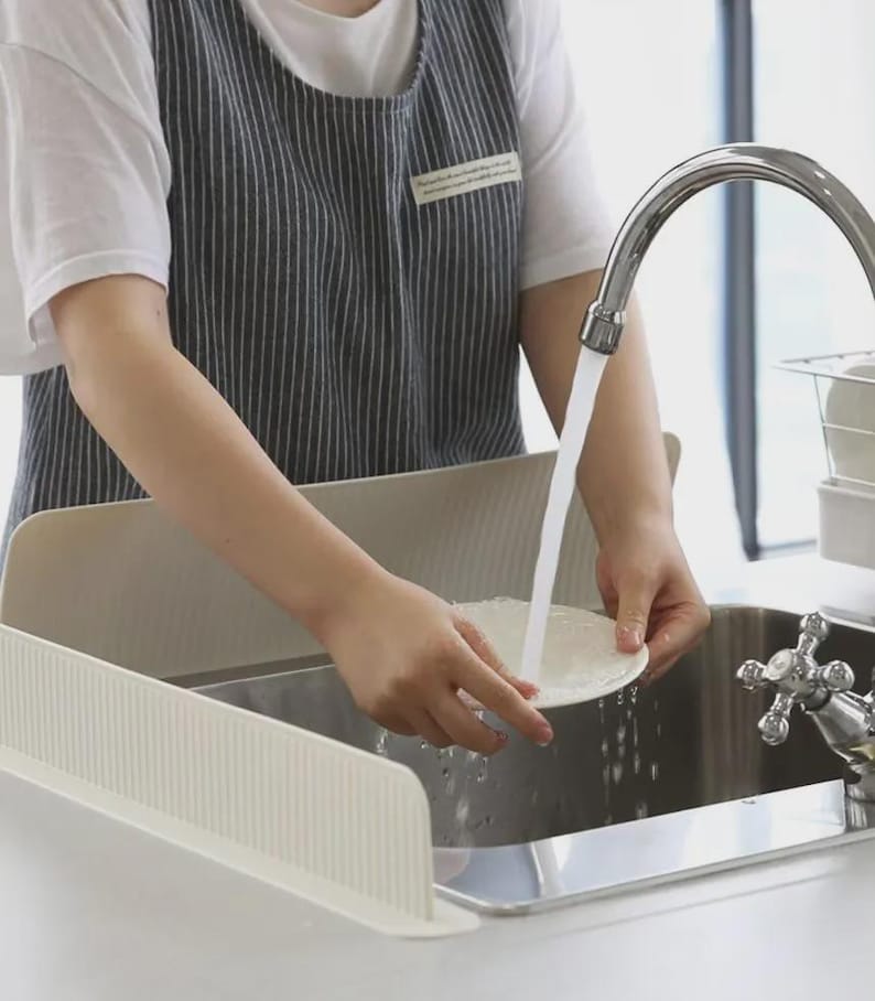 K&ouml;nnte beinhalten: Ein beigefarbener, gerippter, L-f&ouml;rmiger Spritzschutz f&uuml;r die K&uuml;chensp&uuml;le aus flexiblem Material, der am Rand eines Edelstahlbeckens angebracht ist, um beim Abwaschen eines wei&szlig;en Tellers unter dem Wasserhahn Spritzwasser auf die Arbeitsplatte zu verhindern.