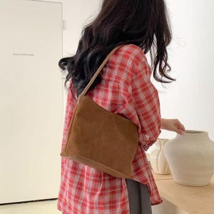 May include: A brown suede shoulder bag with a rectangular shape and a thin strap. The bag is worn over a red and white plaid shirt and gray pants. The background includes a white door and decorative vases.