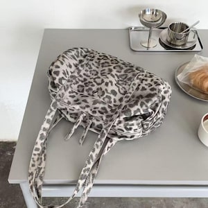 Puede incluir: Un bolso tote grande con un estampado de leopardo en blanco y negro. El bolso tiene dos correas largas y un cierre de cremallera. El bolso est&aacute; sobre una mesa gris.