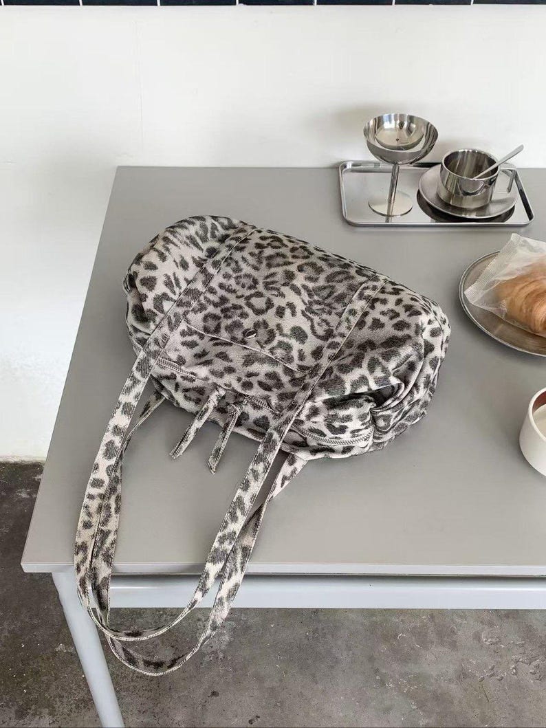 Puede incluir: Un bolso tote grande con un estampado de leopardo en blanco y negro. El bolso tiene dos correas largas y un cierre de cremallera. El bolso est&aacute; sobre una mesa gris.